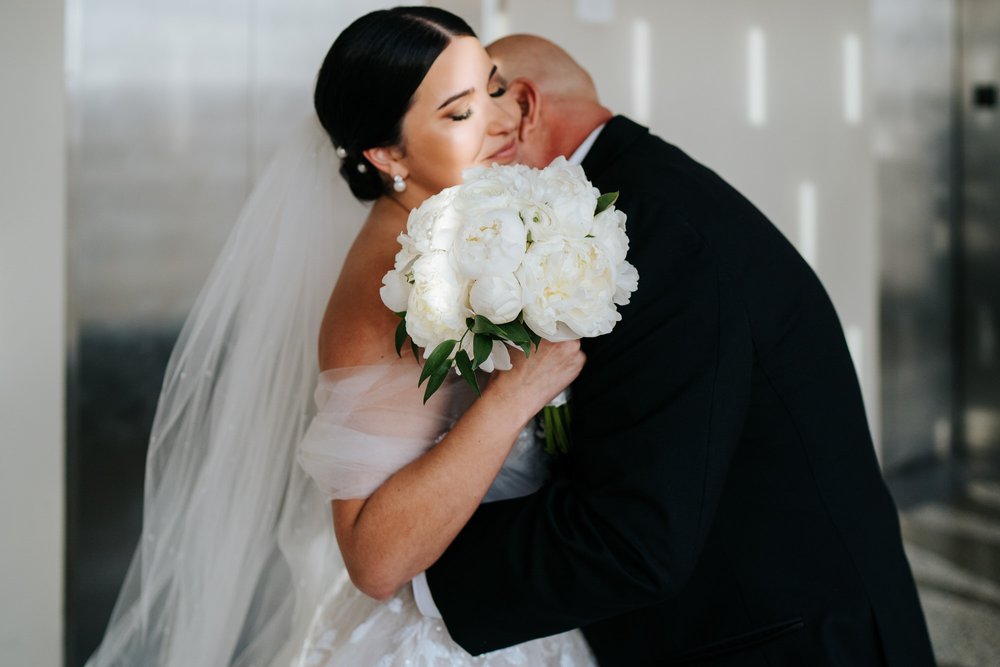 Bride, with a sliver of sunlight on her face, hugs her father who has just seen her in her wedding dress for the first time