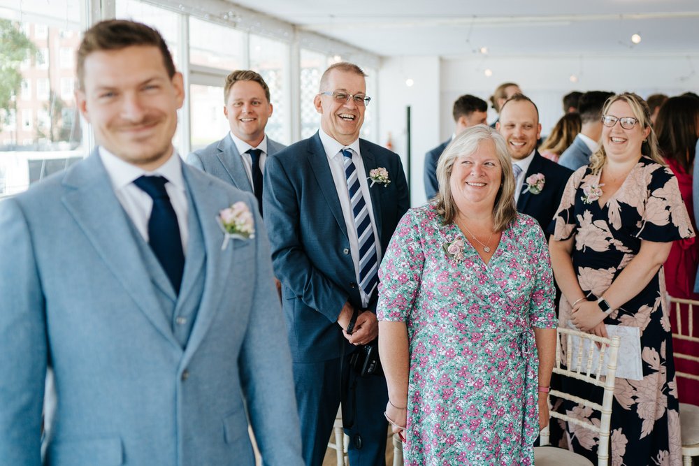 Groom, to the left and out of focus, smirks at camera while his family, in the background, look at him with a coy smile seconds before bride walks down the aisle