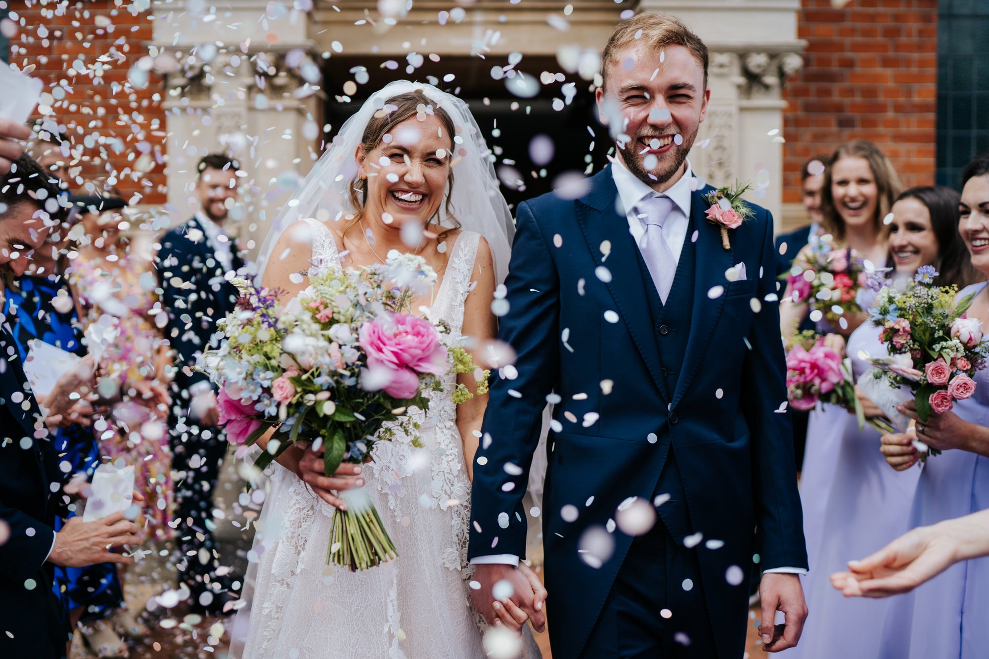 Bride and groom exit church in Wimbledon through tunnel of confetti as they smile in very similar way 