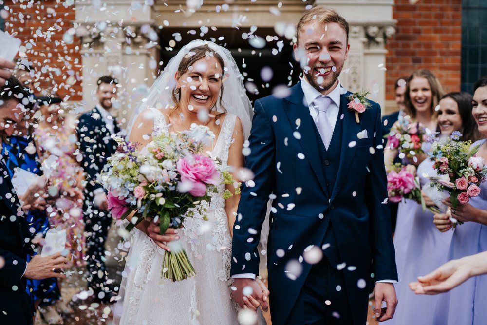 Bride and groom exit church in Wimbledon through tunnel of confetti as they smile in very similar way 