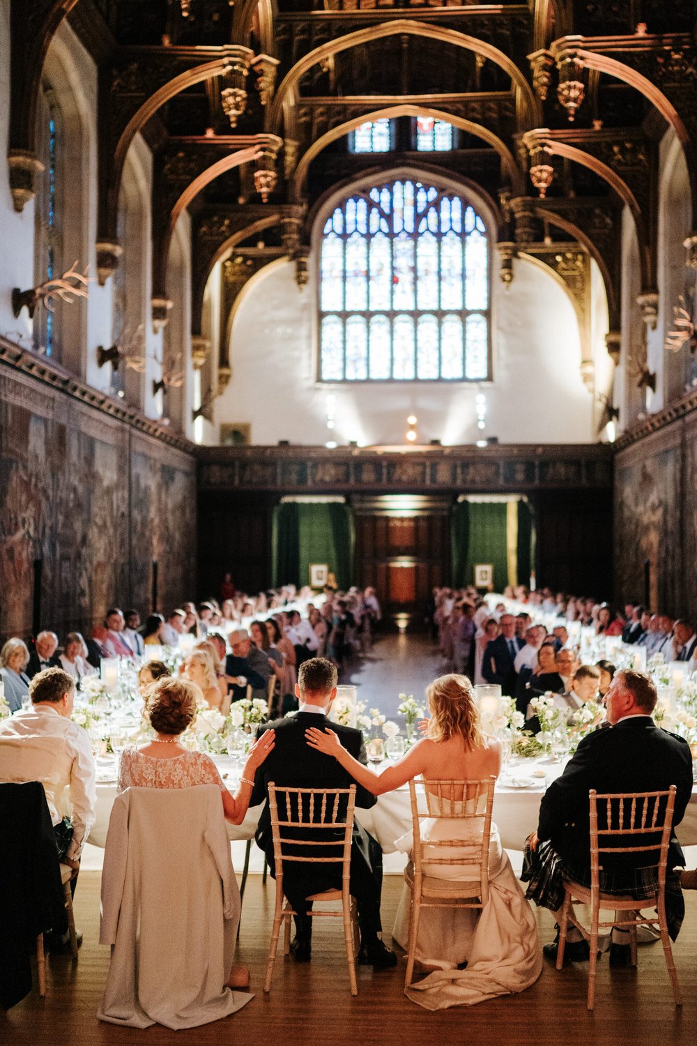 Epic Hampton Court Palace Great Hall is the backdrop for touching photo as groom finishes his speech and both bride and mother of the groom's hand rest on his back 