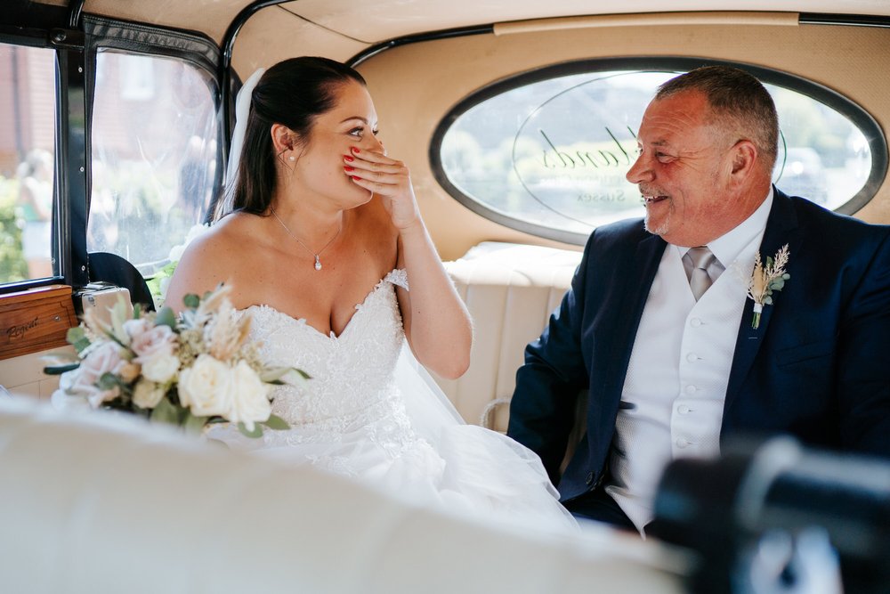 Bride, covering her mouth out of excitement, looks at her father as they are both sitting in the back of the wedding car, minutes before the ceremony begins