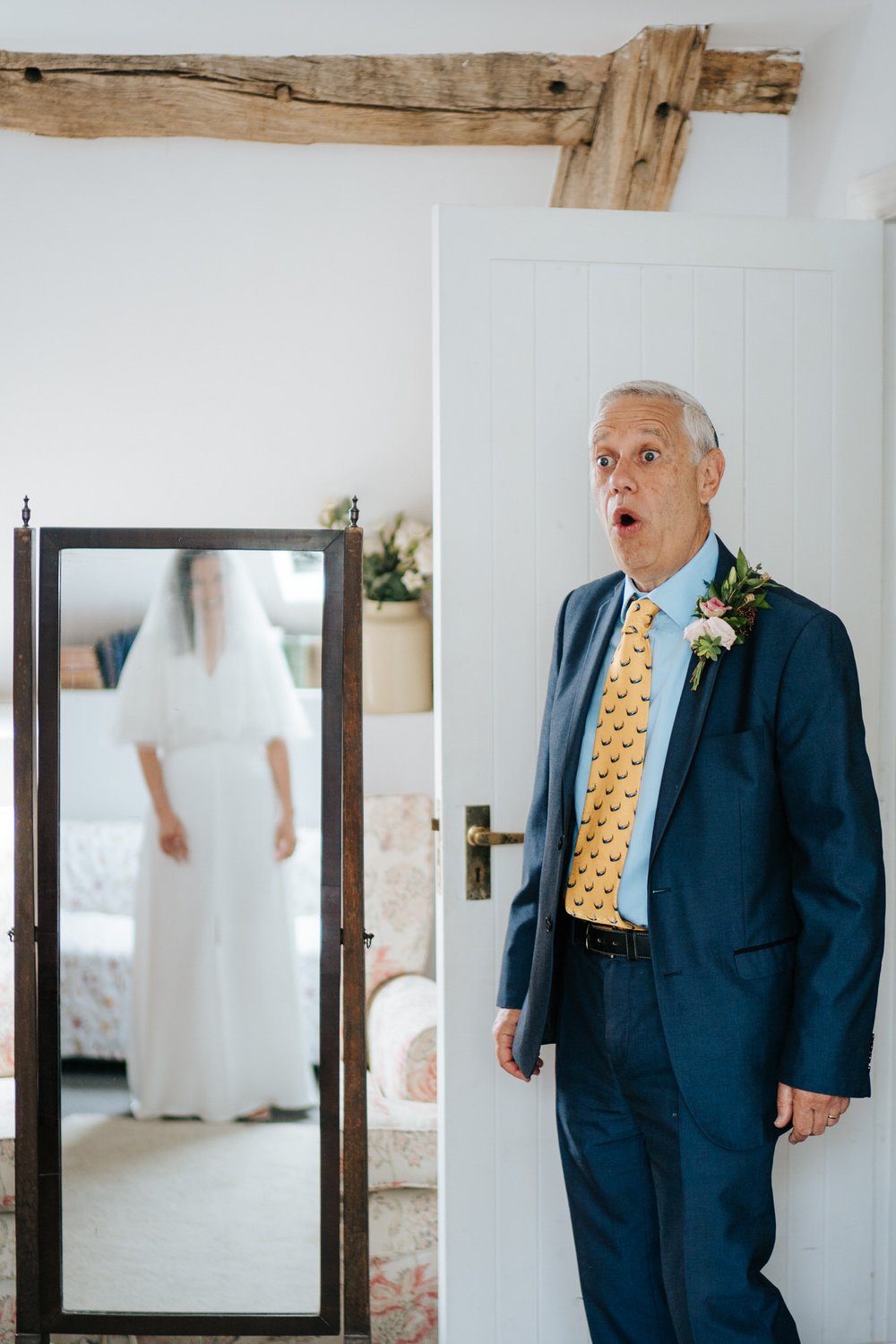 Father of the bride, with jaw on the floor, sees his daughter in her dress for the first time as the daughter can be seen, out of focus, in mirror reflection