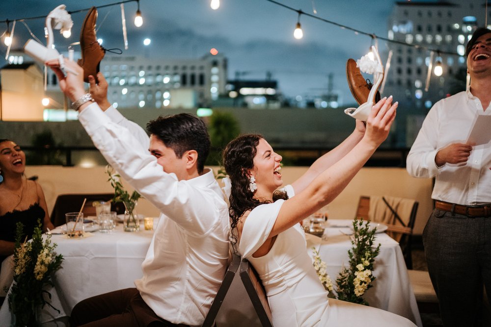 Bride and groom, flanked by their best man and maid of honour, hold up each other's shoes as they play funny game during rehearsal dinner celebration