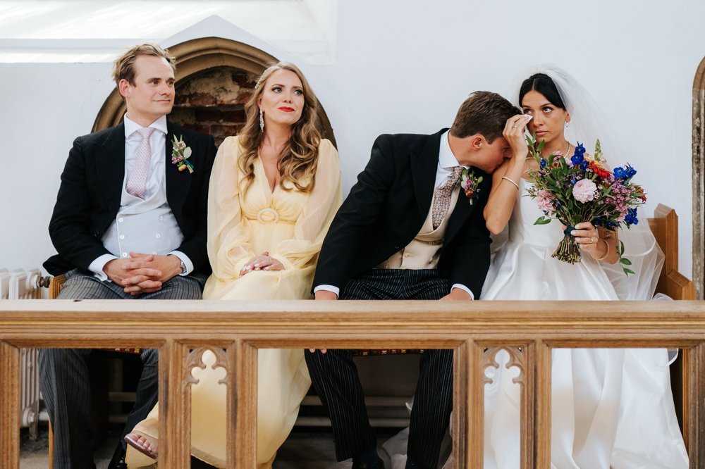 Bride wipes her tears as groom gives her a kiss on the shoulder during wedding day church ceremony as guests sit next to them