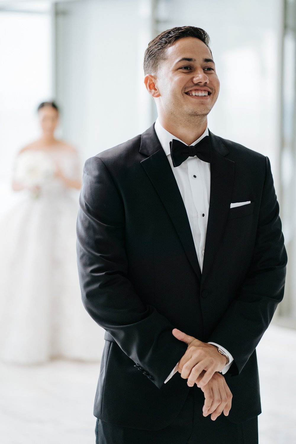 Groom, in foreground, stands excitedly with hands clasped as bride, out of focus, starts approaching him for the first look
