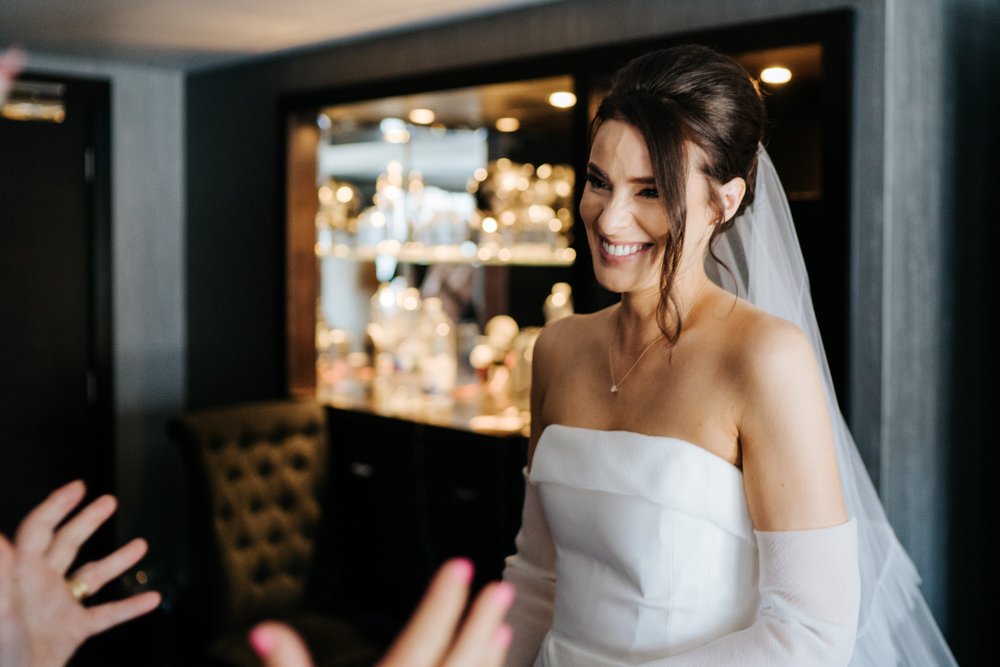 Bride, in her wedding dress, smiles as her mum, off camera, stretches her hands out to embrace her