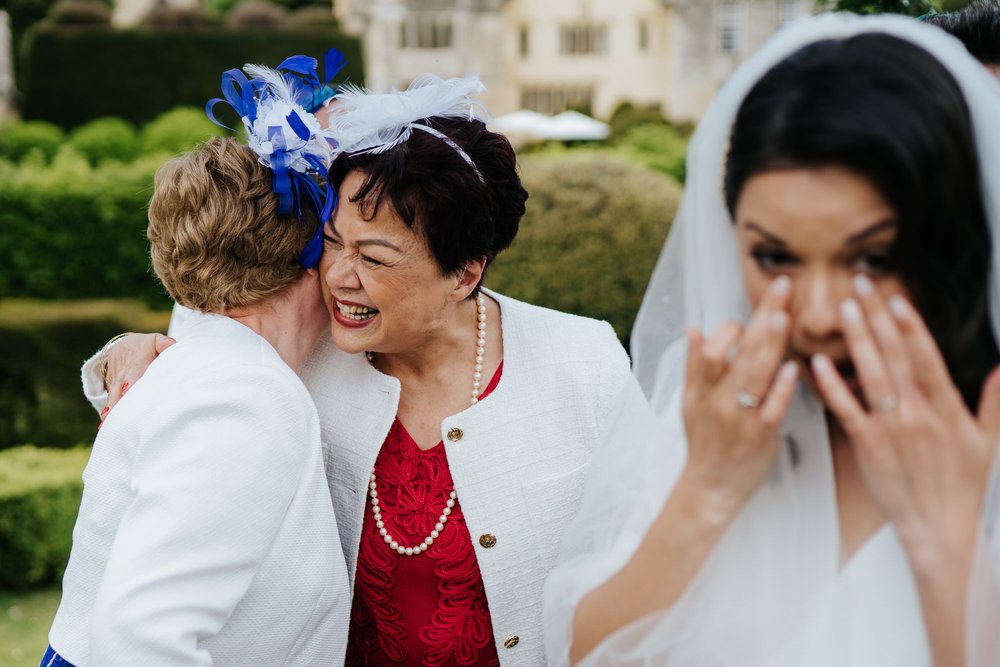 Bride, out of focus and in the front, wipes her tears as mother of the bride and mother of the groom hug each other in the background