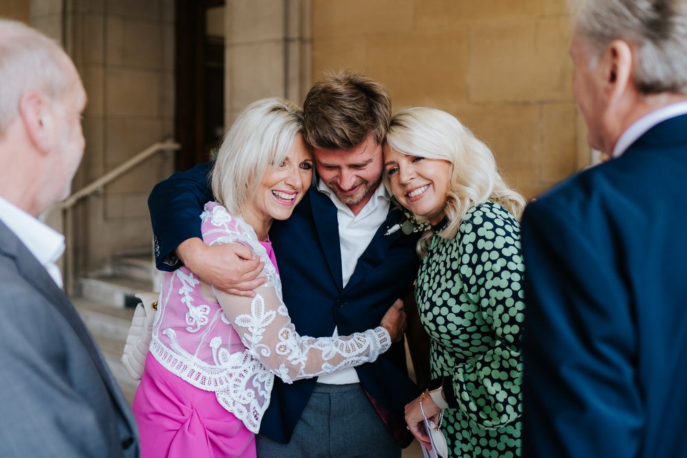 Groom hugs his mother and his sister in the middle of the frame as he is flanked by his father and the father of the bride, both out of focus