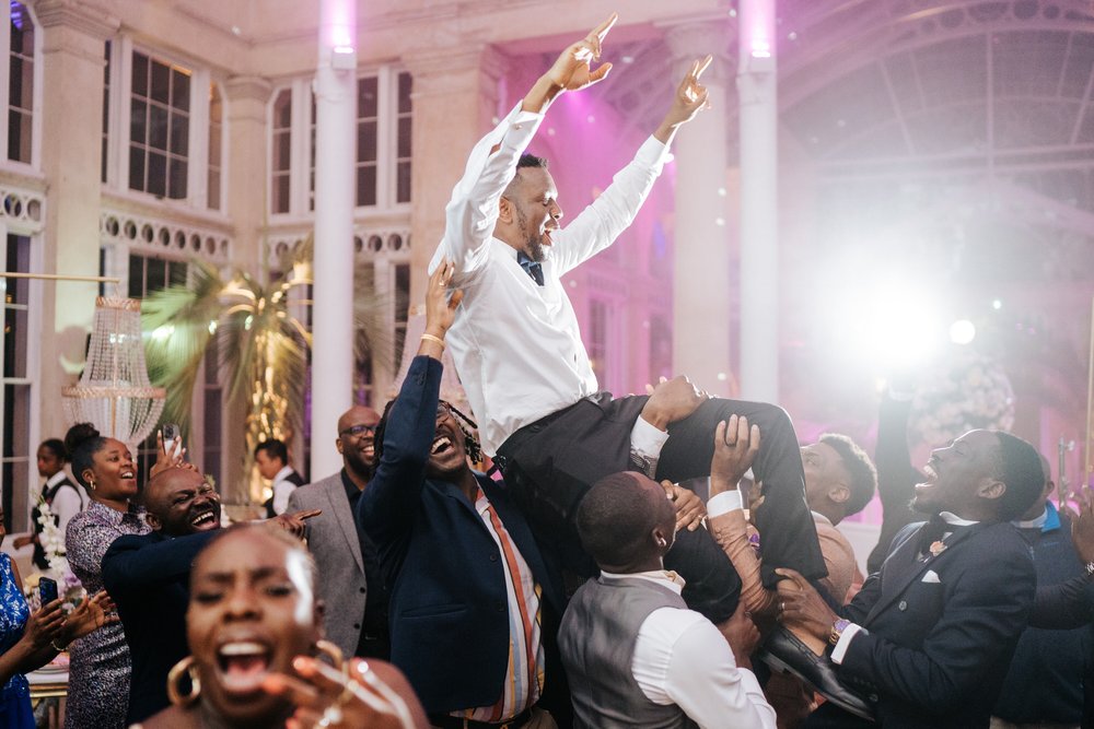 Groom throws his hands in the air as he is held up by his friends during Nigerian wedding day celebration at Syon Park