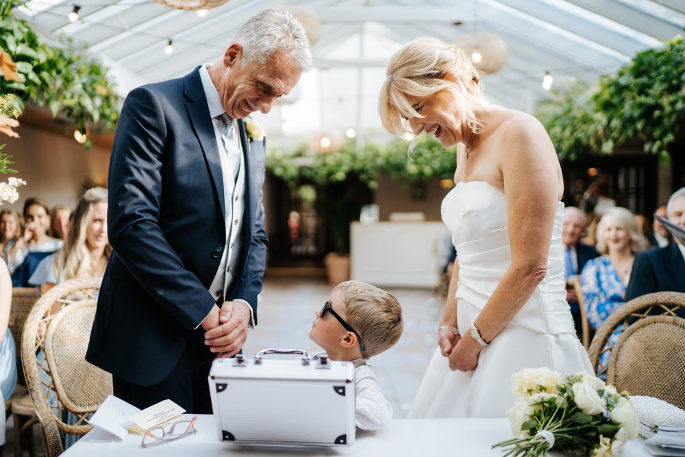 Bride and groom look down at ring bearer, dressed in sunglasses and with a briefcase, as the young boy hands them both their wedding bands