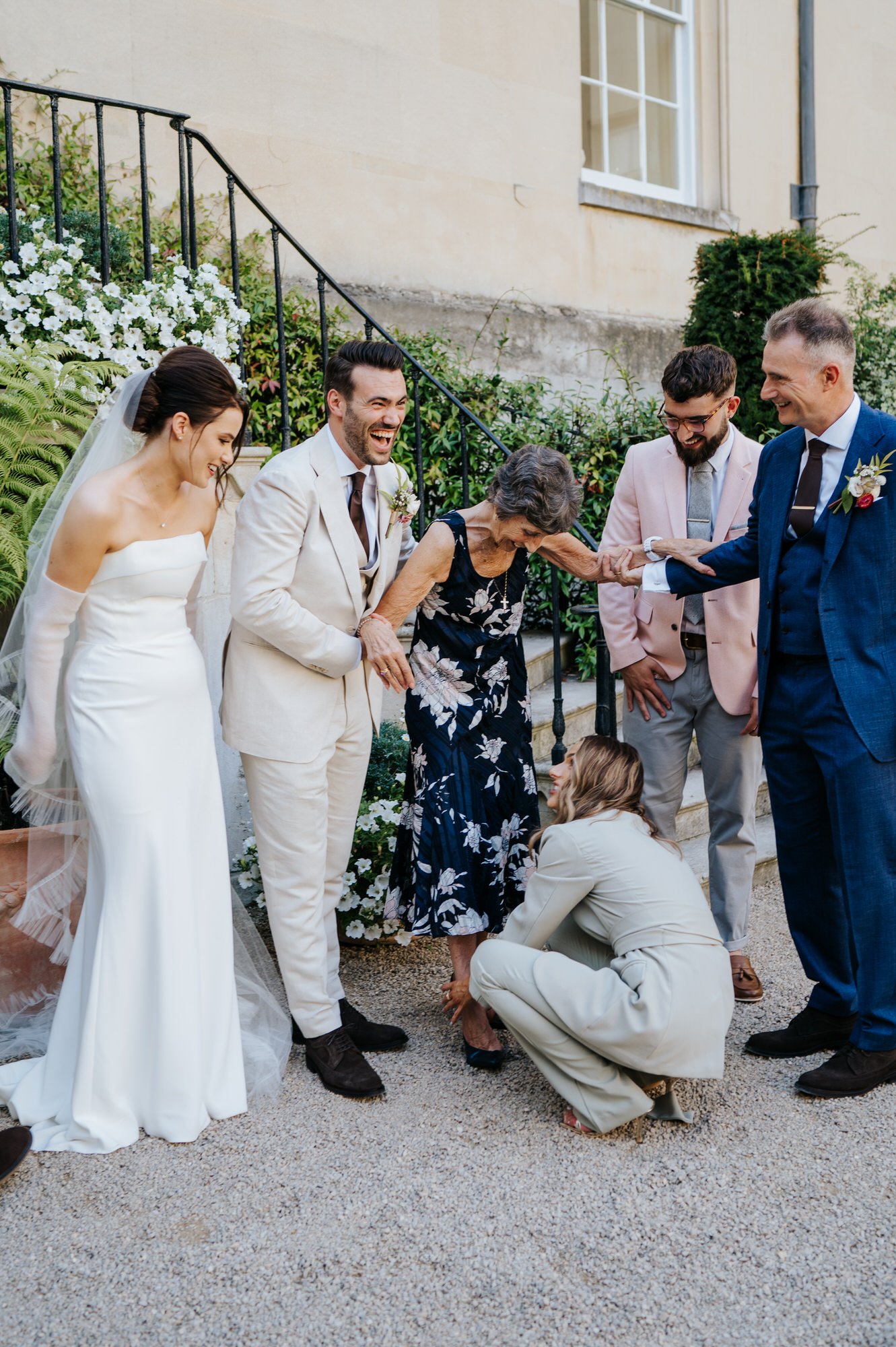 Groom in hysterics after funny moment while groom's sister helps the grandmother into her shoes during formal family photos at Syon Park
