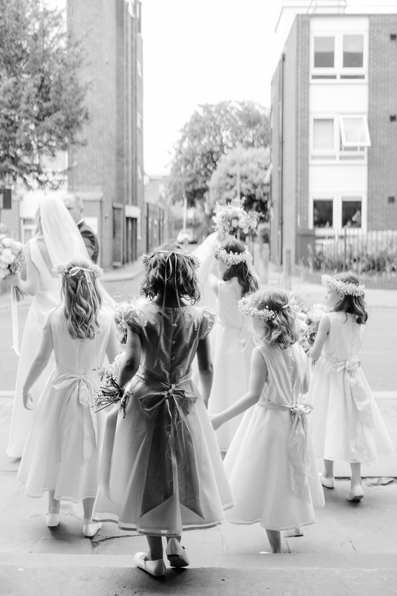 Black and white image of five little bridesmaids helping the bride exit the church as they hold her dress from behind
