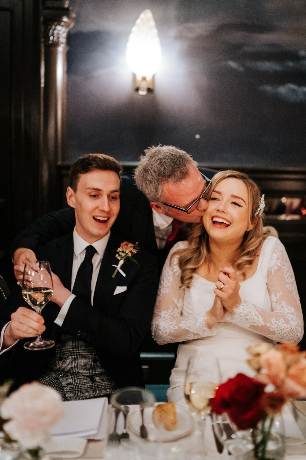 Father of the groom kisses bride while embracing his son after he delivered his wedding speech at Nomad Hotel in London