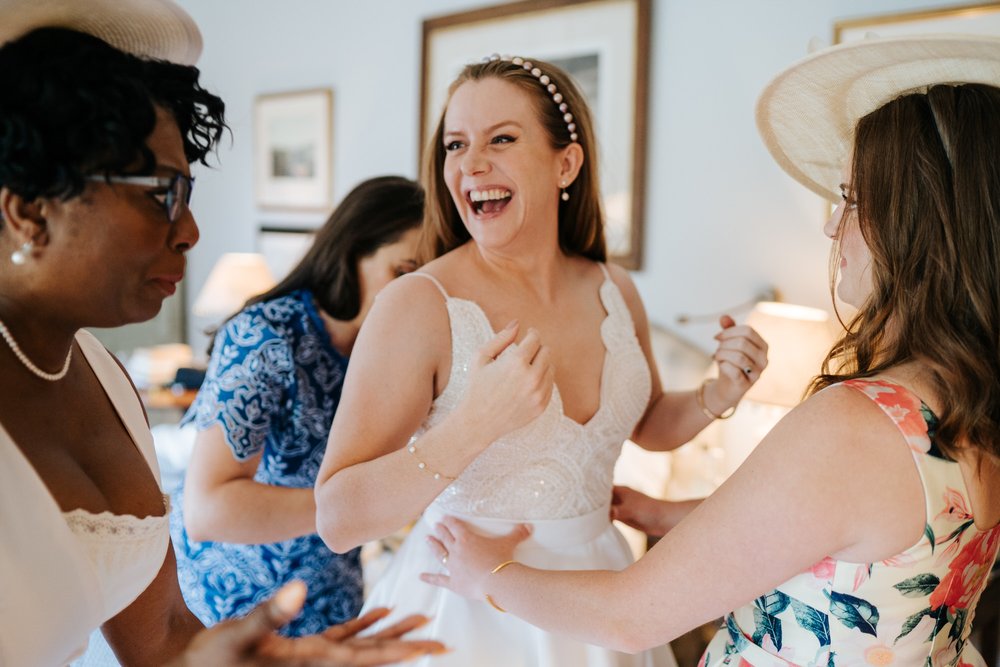 Bride smiles as three of her friends help her get into her wedding dress while one has a very panicked look on her face