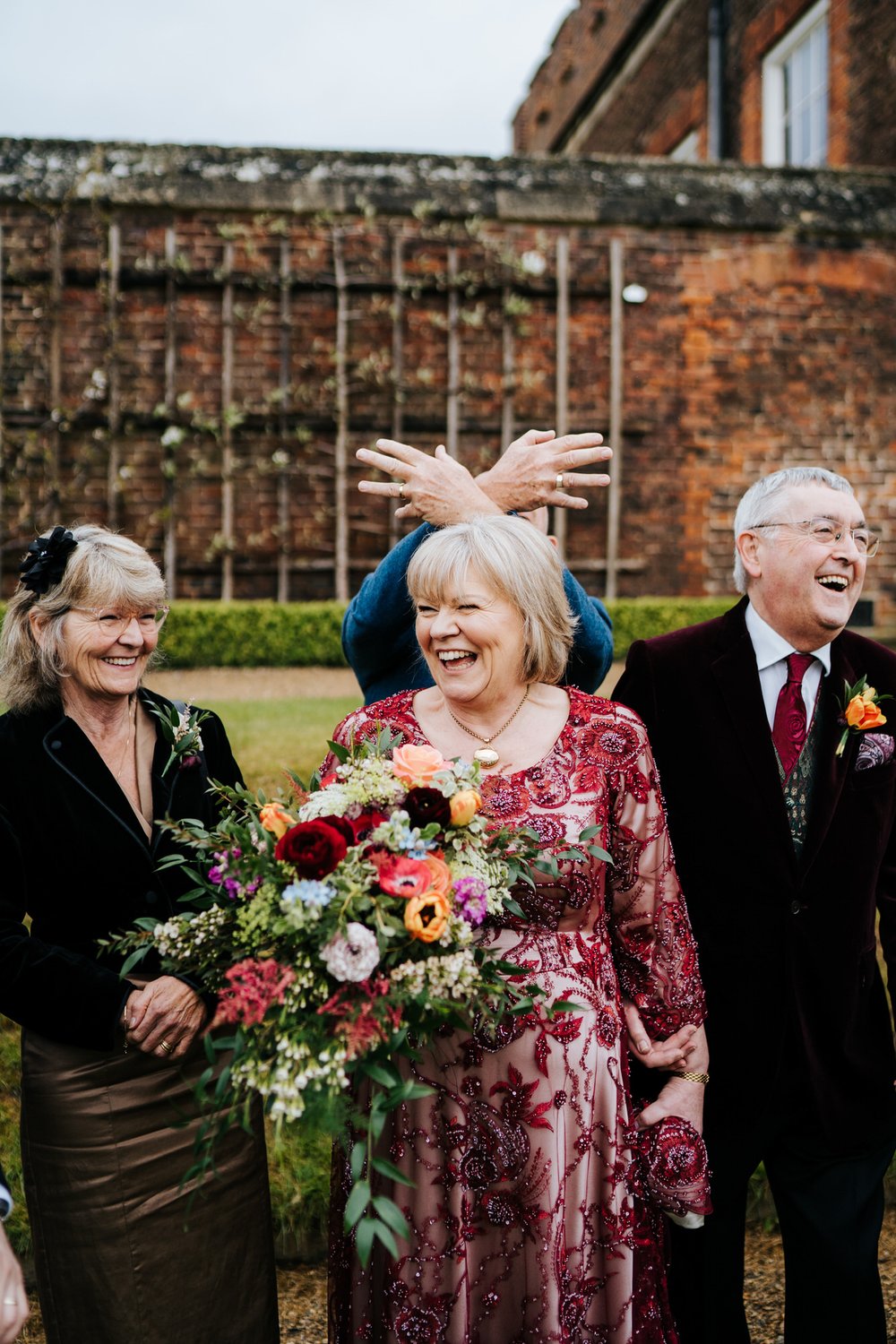 Bride, flanked by groom and sister, stands and holds her beautiful bouquet as someone makes a silly hand sign on top of her head