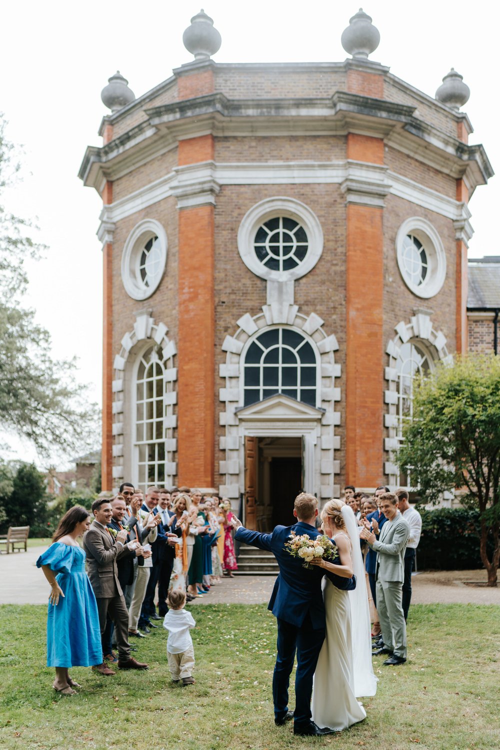 Photo of bride and groom waving to all of their guests after they finish walking through the confetti tunnel, with Orleans House Gallery in the background
