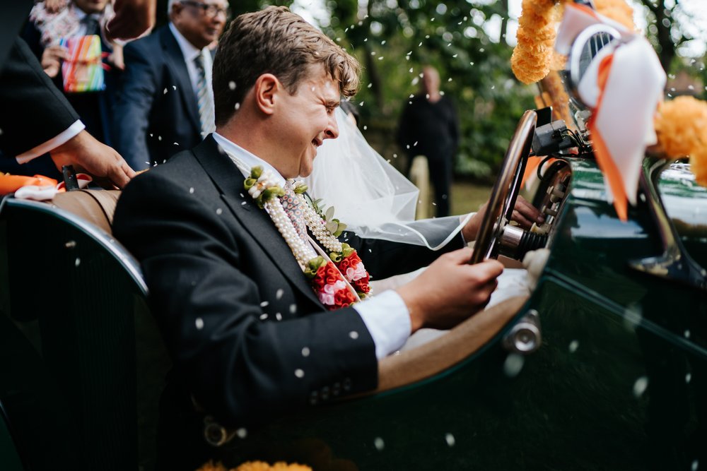 Close-up photograph of groom in his new vintage wedding car as he is pelted by rice