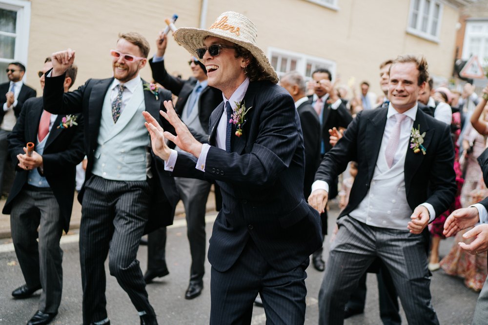 Groomsman wearing tropical hat dances on the streets of Woodbridge during wedding day Baraat