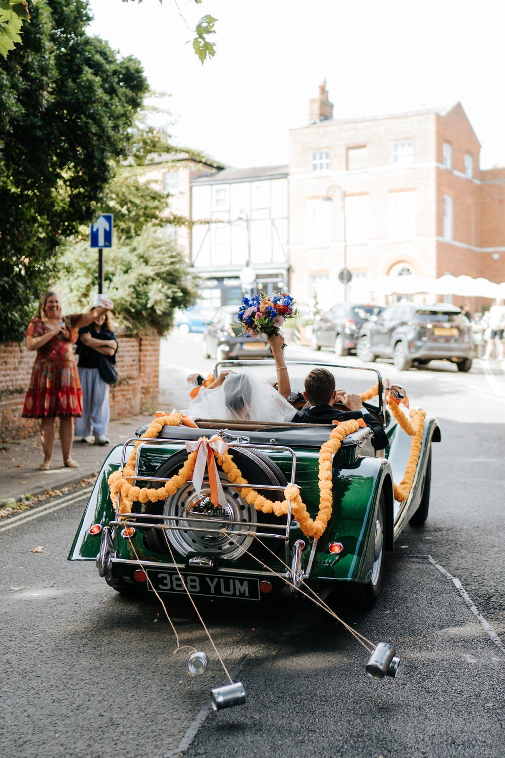 Bride and groom drive away in vintage Morgan as bride, in passenger seat, lifts her bouquet in celebration