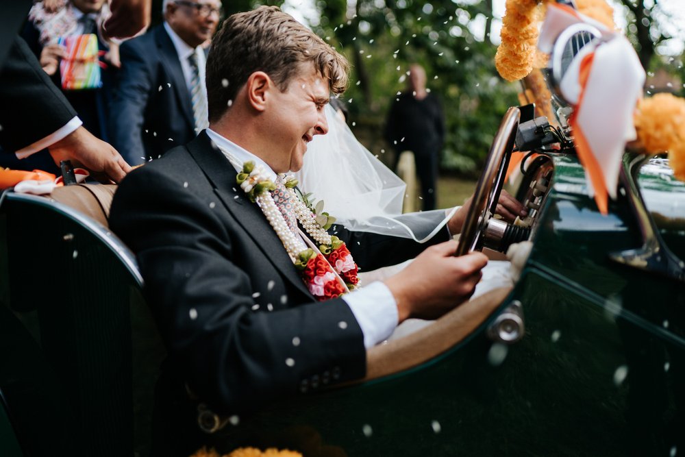 Groom, sitting in his vintage Morgan car, is pelted with rice as part of wedding tradition