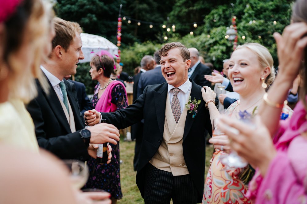 Candid photograph of groom talking to guests and laughing with them during garden marquee drinks and canapes