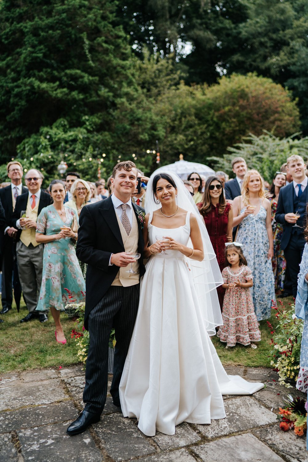 Bride and groom, hand in hand, listen to the father of the bride speech while standing in the garden