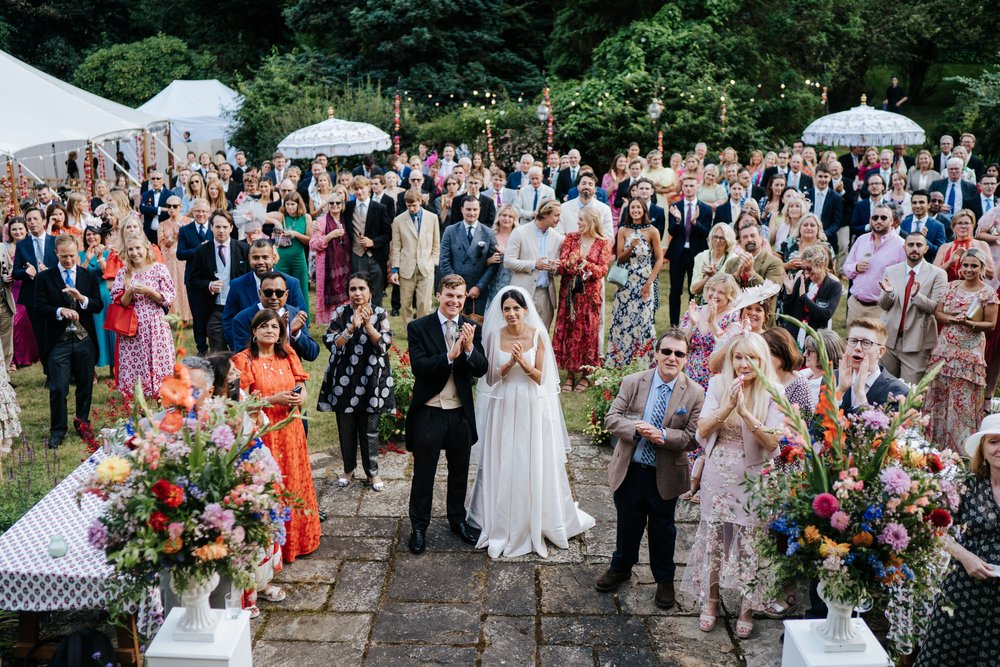 Wide photograph of bride and groom, front and center, and guests clapping as the father of the bride finishes his wedding speech in beautifully lush English garden