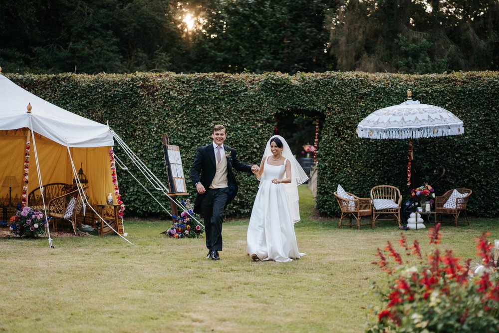 Bride and groom make an entrance into their marquee as the sun pokes through the trees that can be seen behind them