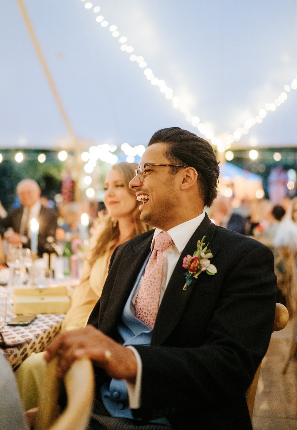 Bride's brother smiles radiantly during wedding speeches taking place inside atmospherically-lit marquee at British garden marquee wedding