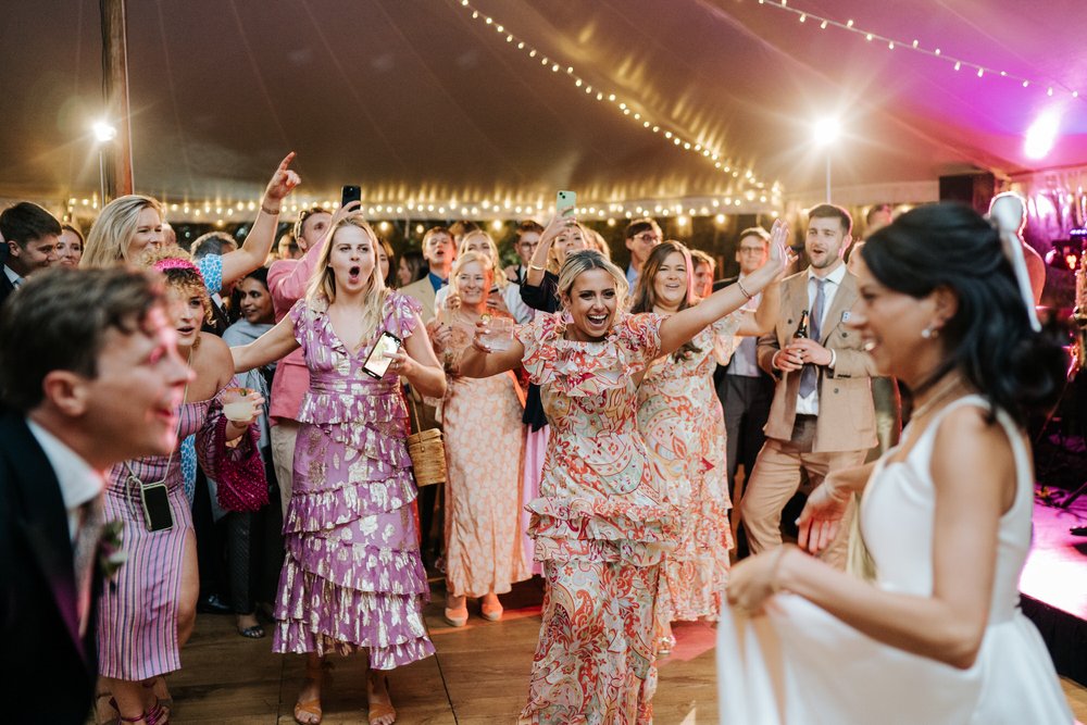 Bride and groom, front and centre but out of focus, look at each other as other guests on the dancefloor cheer them on