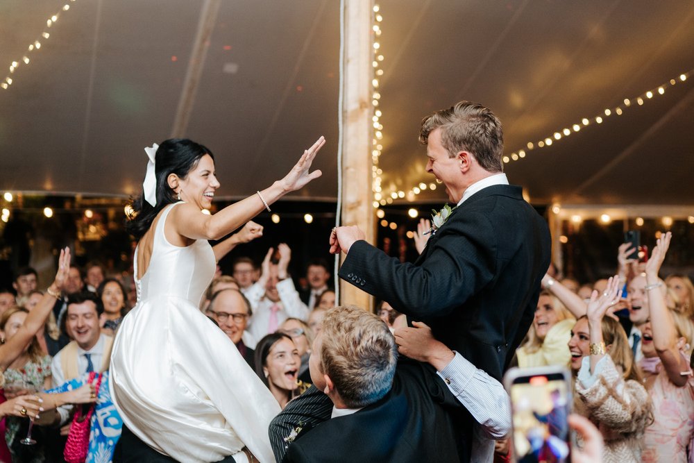 Bride and groom are hoisted up on guests's shoulders as they throw their hands in the air and dance during wedding party