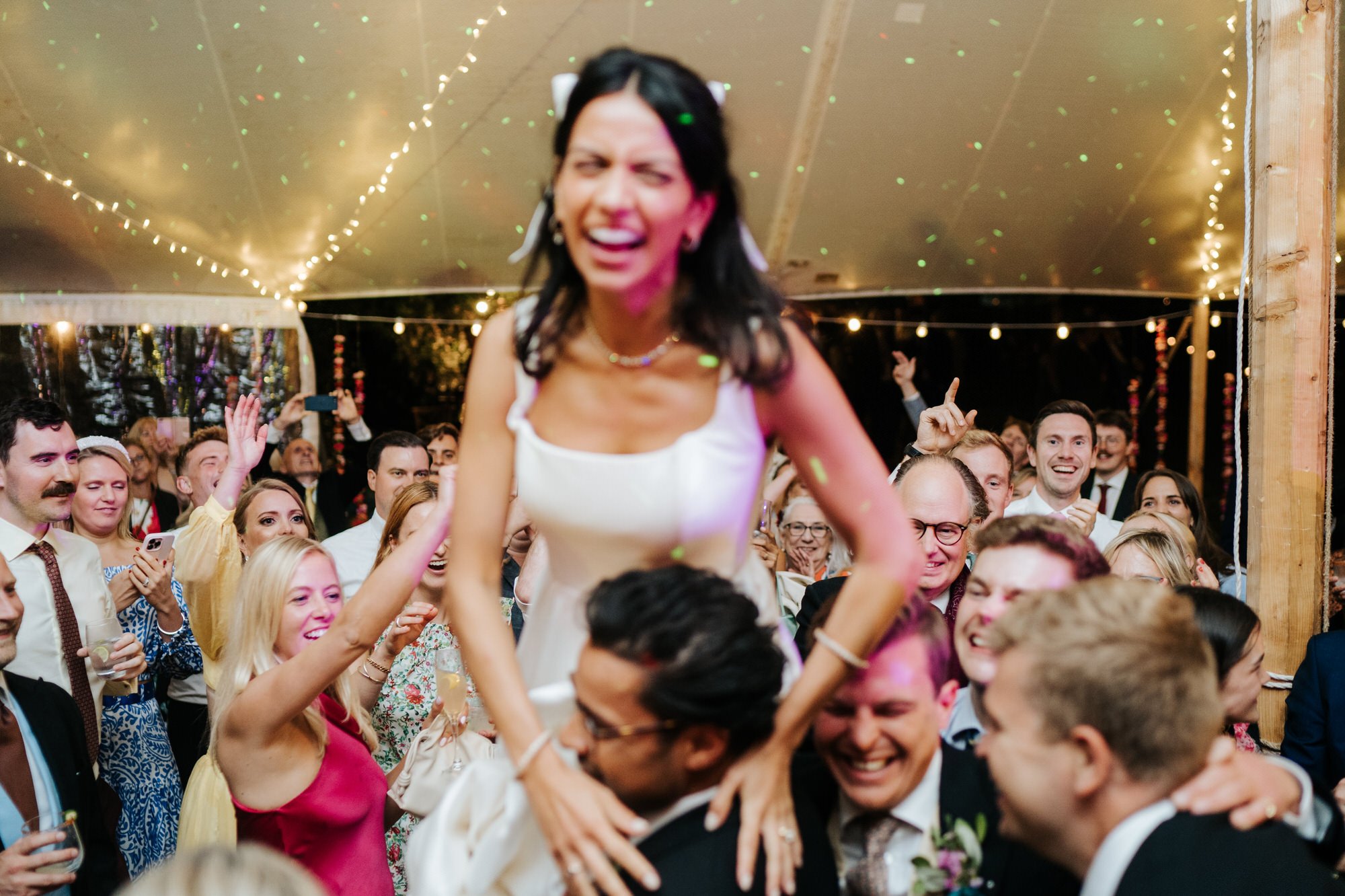 front and centre bride, out of focus, is lighted up and laughs while her guests smile in the background