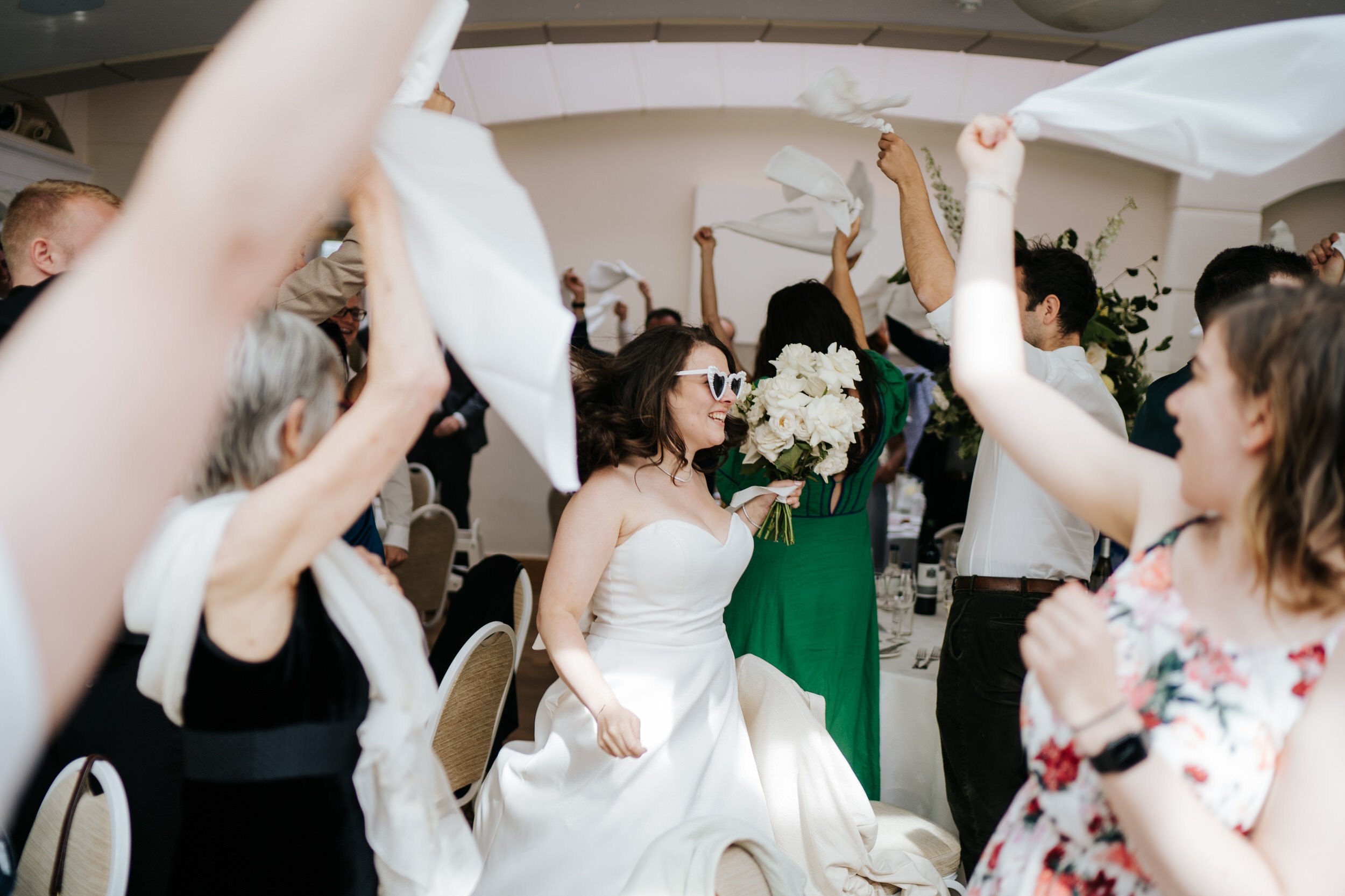 Bride, wearing sunglasses, makes entrance into room at Pembroke Lodge as her guests wave napkins in the air