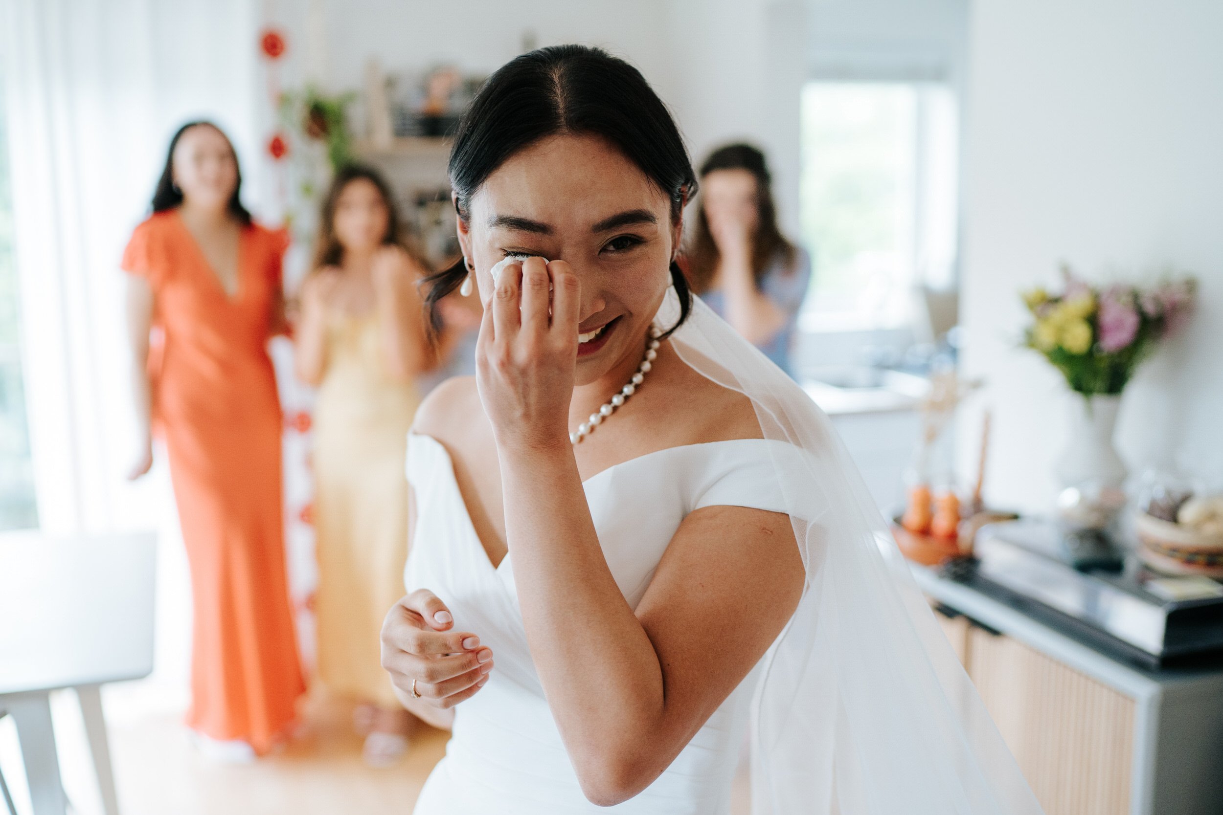 Bride, turned to camera, wipes tear of her face as her bridesmaids, out of focus in background, see her in her dress for the first time