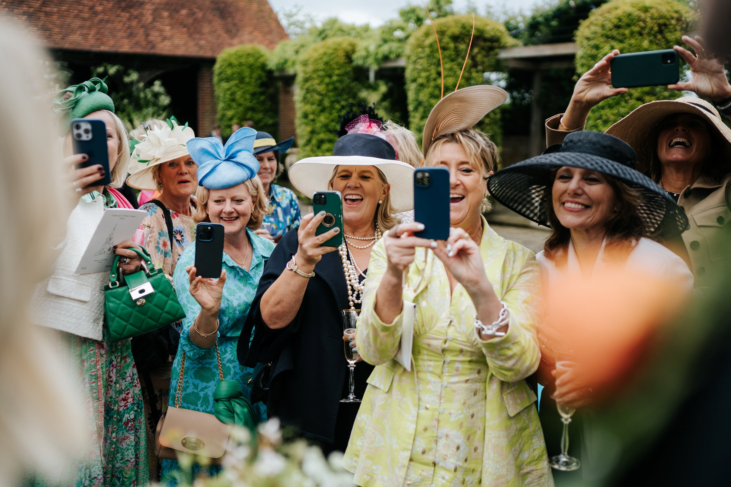 Group of women wearing fancy hats all smile and take photos of bride standing off the side of the frame