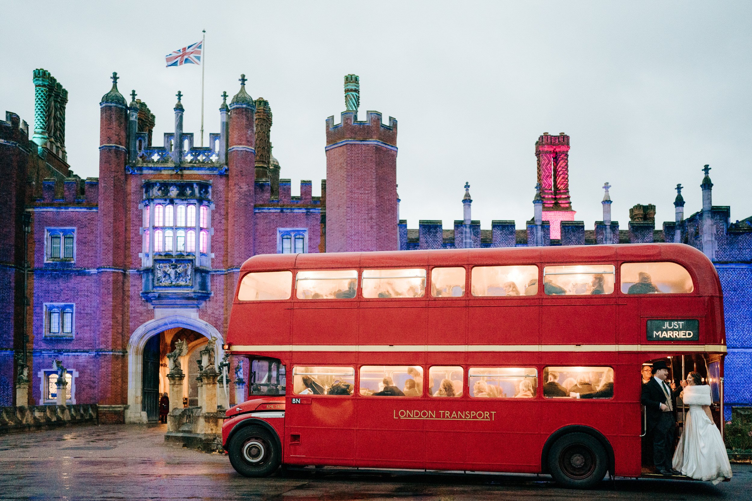 Wide photograph of double-decker London bus arriving at Hampton Court Palace during gloomy winter day as bride and groom stand in the back entrance