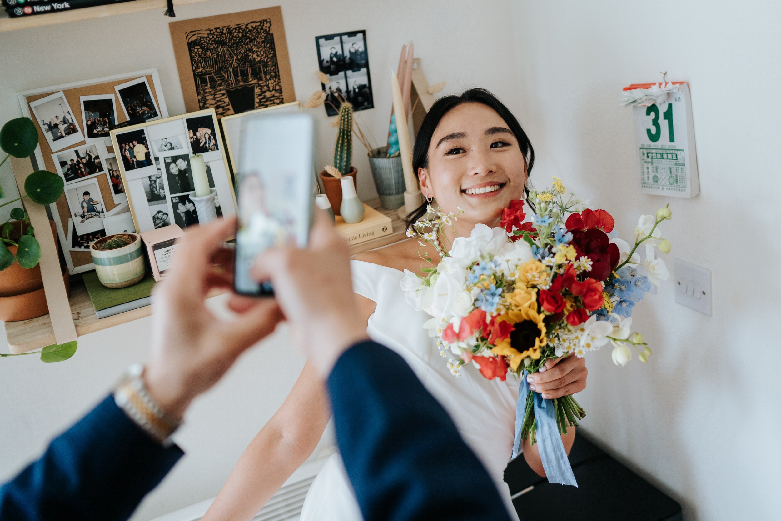 Bride poses in front of calendar showing the day's date as her father takes a photo on his phone