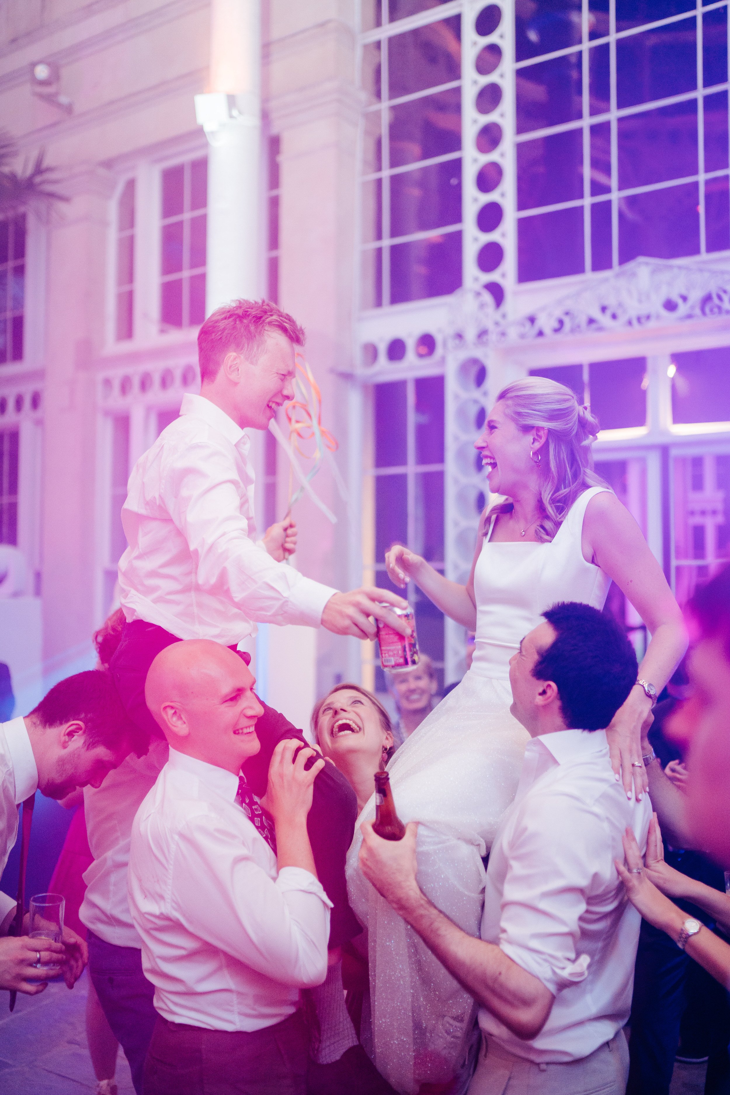 Bride and groom having a blast during wedding party as they sit on friends' shoulders and are drenched in pink and purple light