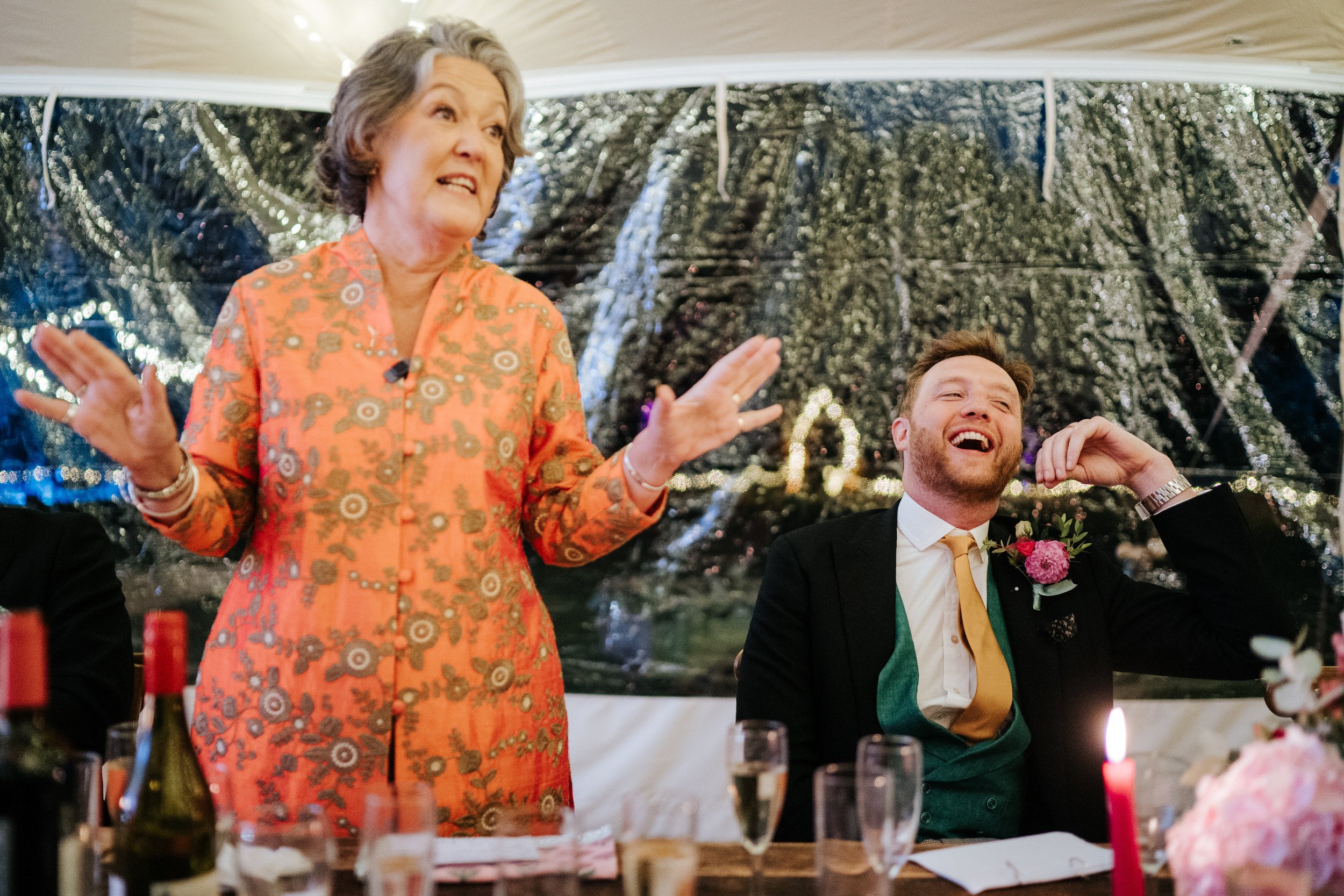 Mother of the bride passionately delivers a wedding speech as the groom, next to her, leans back and laughs in Little Dover House wedding