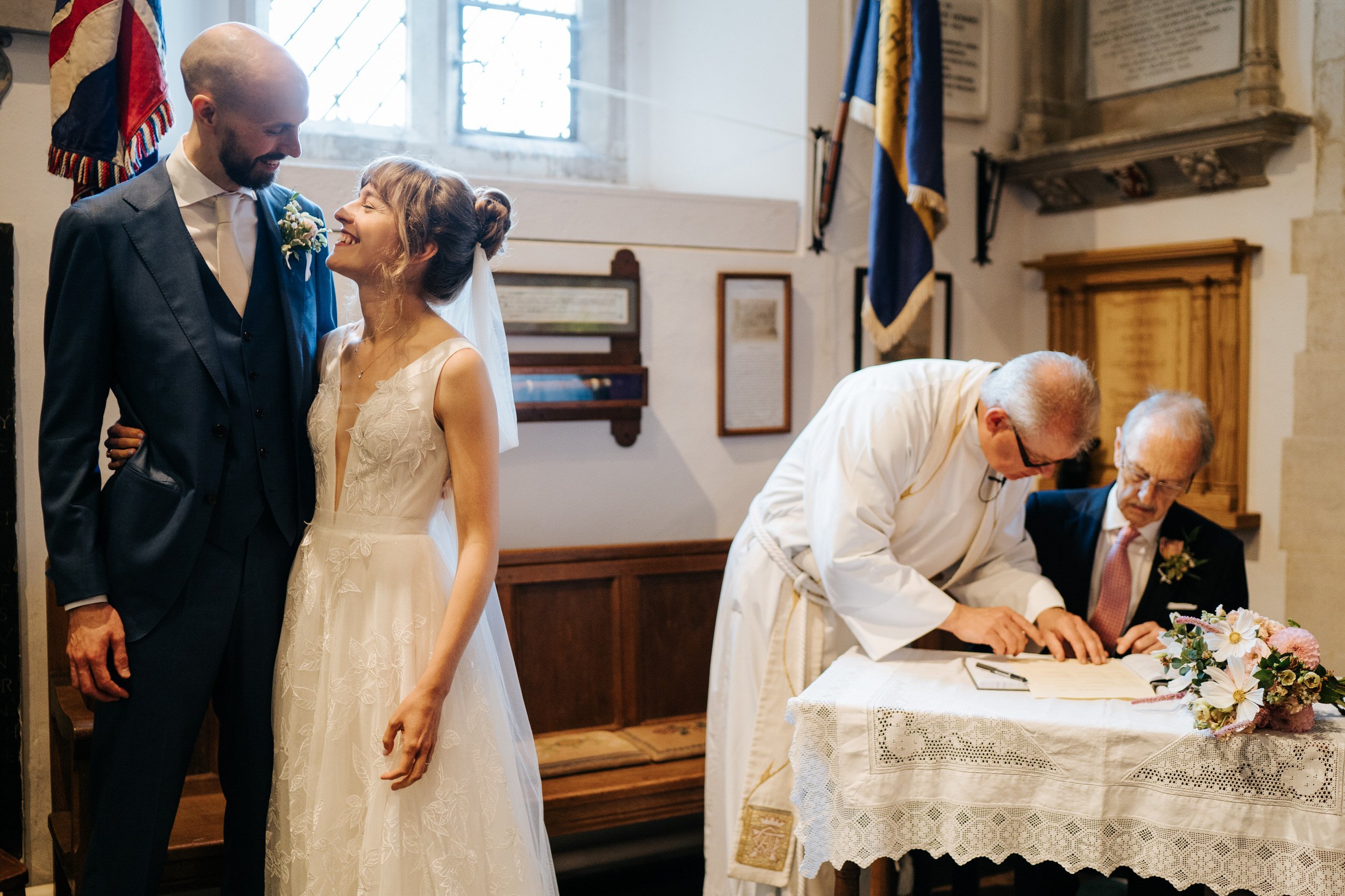 Bride and groom embrace as the vicar and bride's father sign the wedding registry in the background