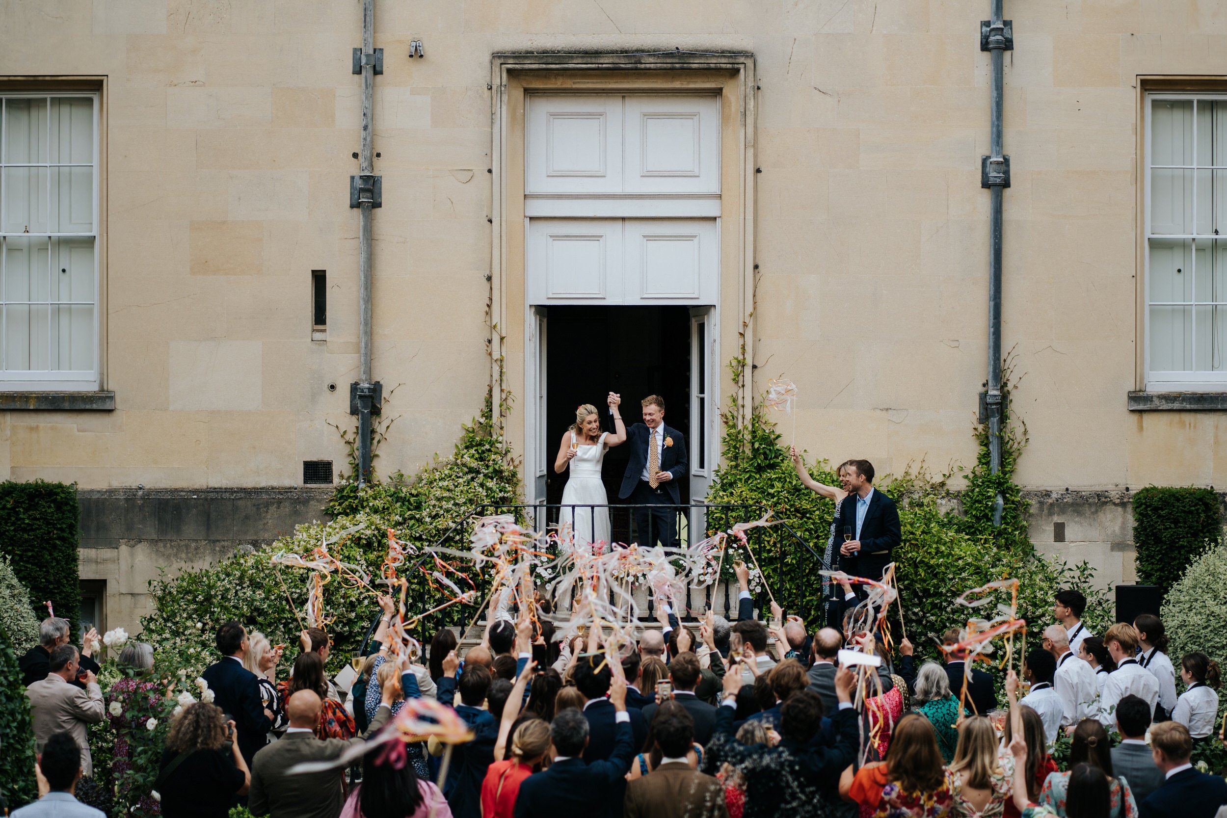 Wide photograph of bride and groom making their entrance into drinks reception at Syon Park as guests wave streamers