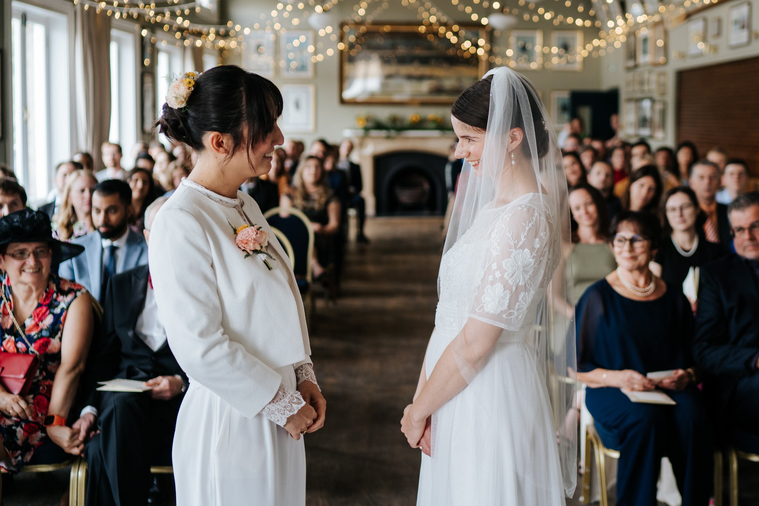 Two brides look at each other lovingly during ceremony at Putney Rowing Club as lights twinkle in the background