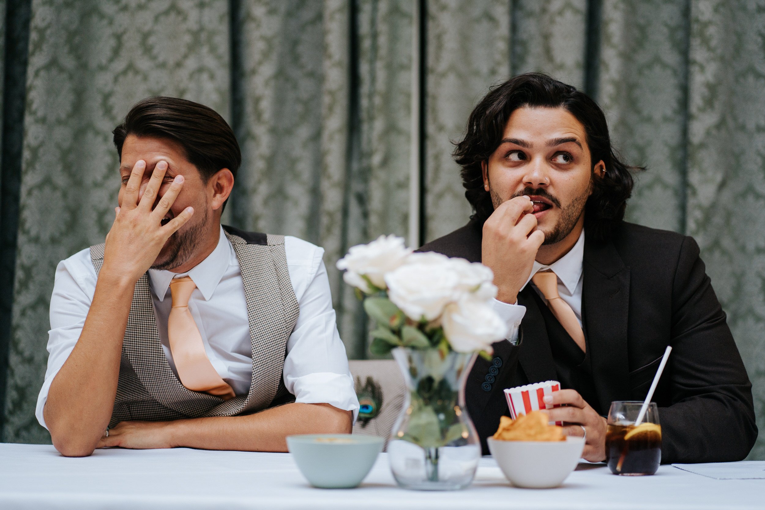 Close-up of two grooms reacting to wedding speeches as one of them covers his face with his hand and the other eats popcorn
