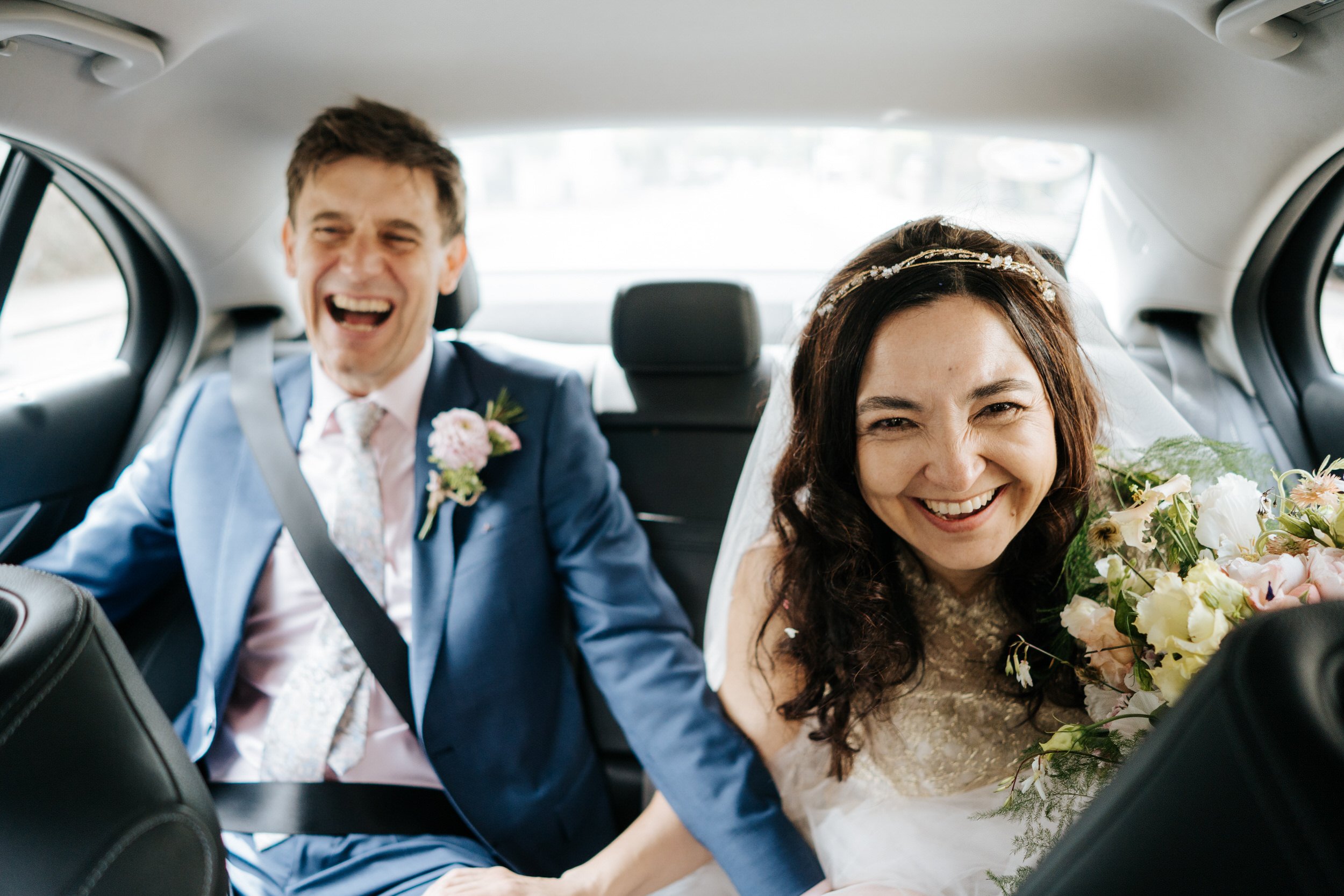 Bride and groom sitting in the back of a car laugh and smile as they make their way from wedding ceremony to wedding reception