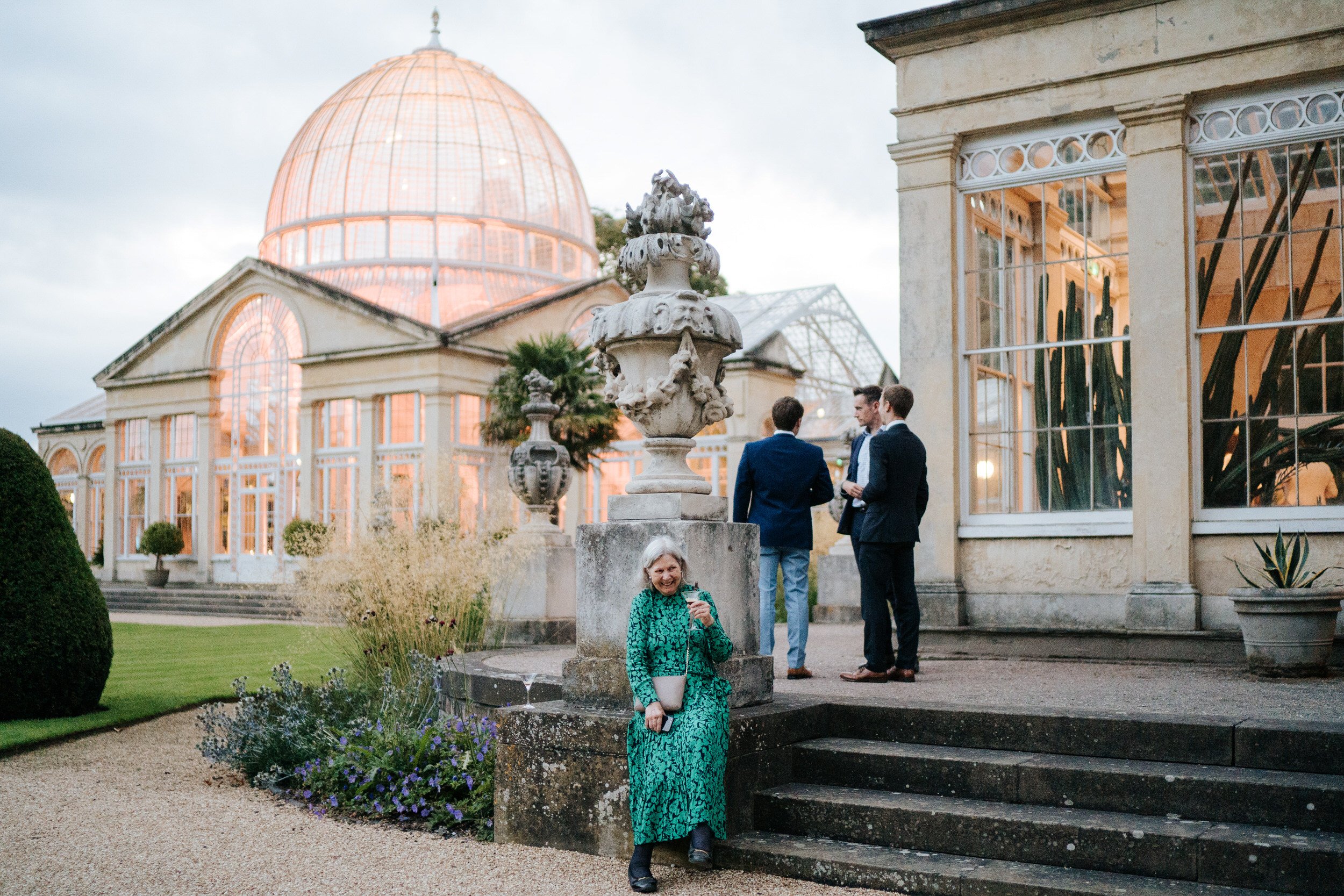 Wide photograph of Syon Park conservatory in the background as woman holds drink and has a cheeky smile while sitting on concrete steps