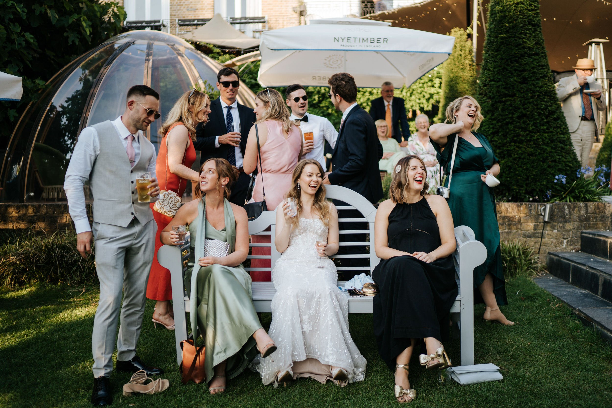 Atmospheric photograph of wedding guests and bride sitting on a bench during sunny afternoon at Bingham Riverhouse
