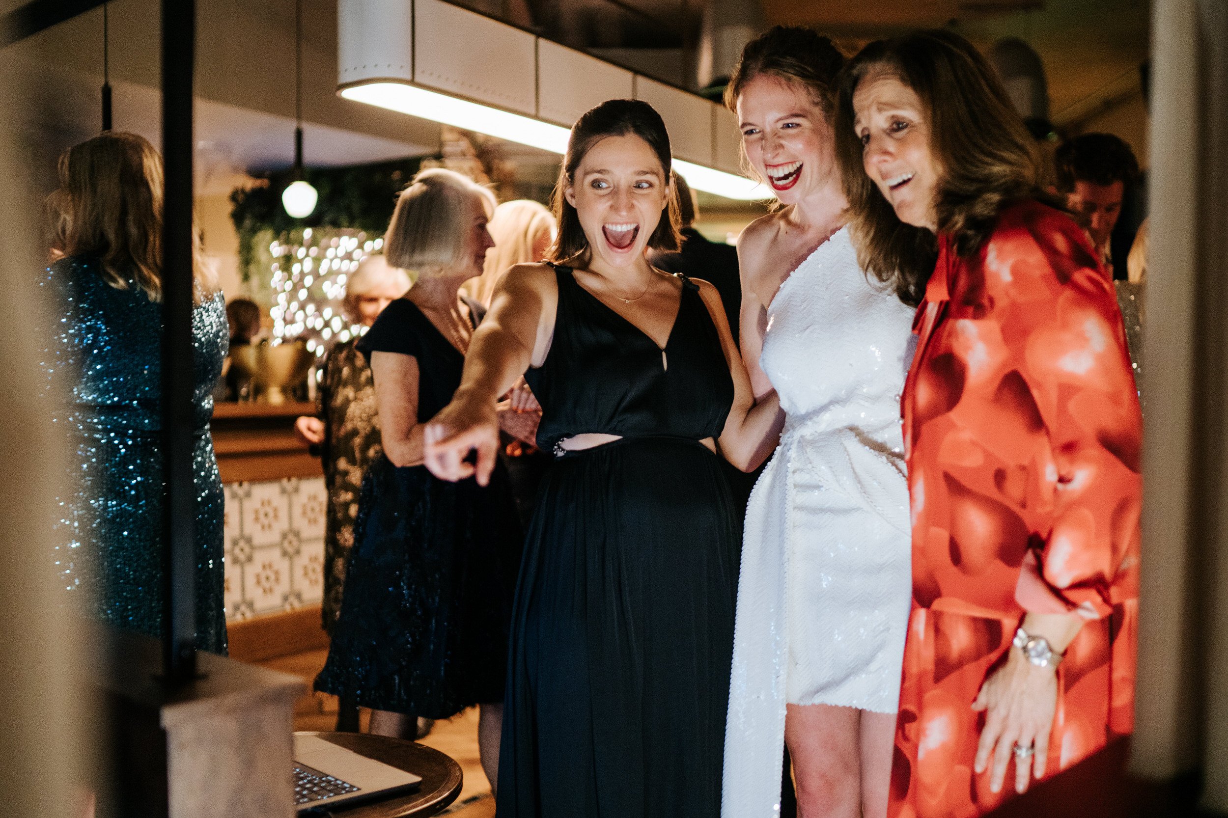 Bride, mother and friend look in excitement at screen showcasing selection of wedding photographs