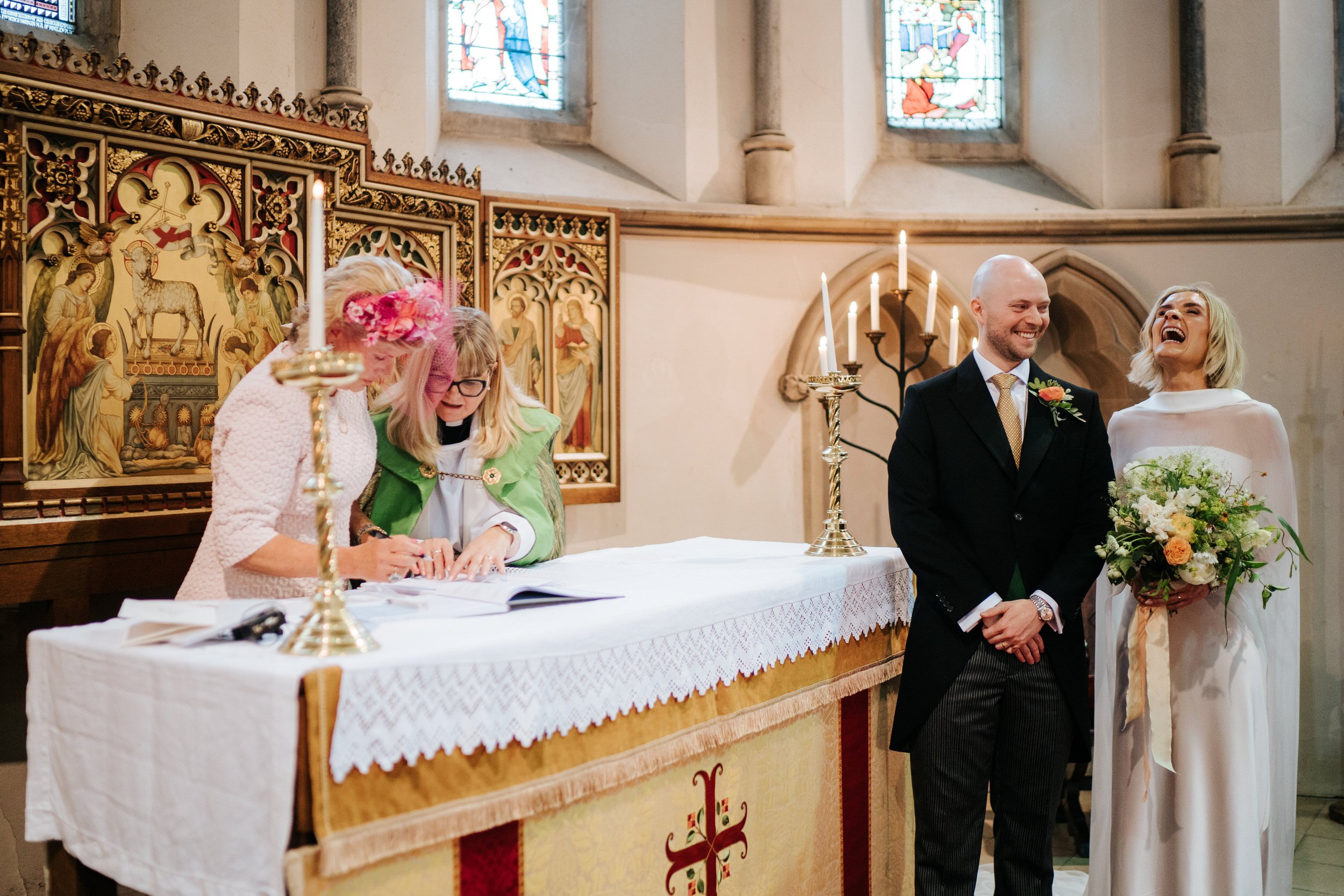 Bride and groom laugh and smile as vicar and bride's mom sign the wedding certificate