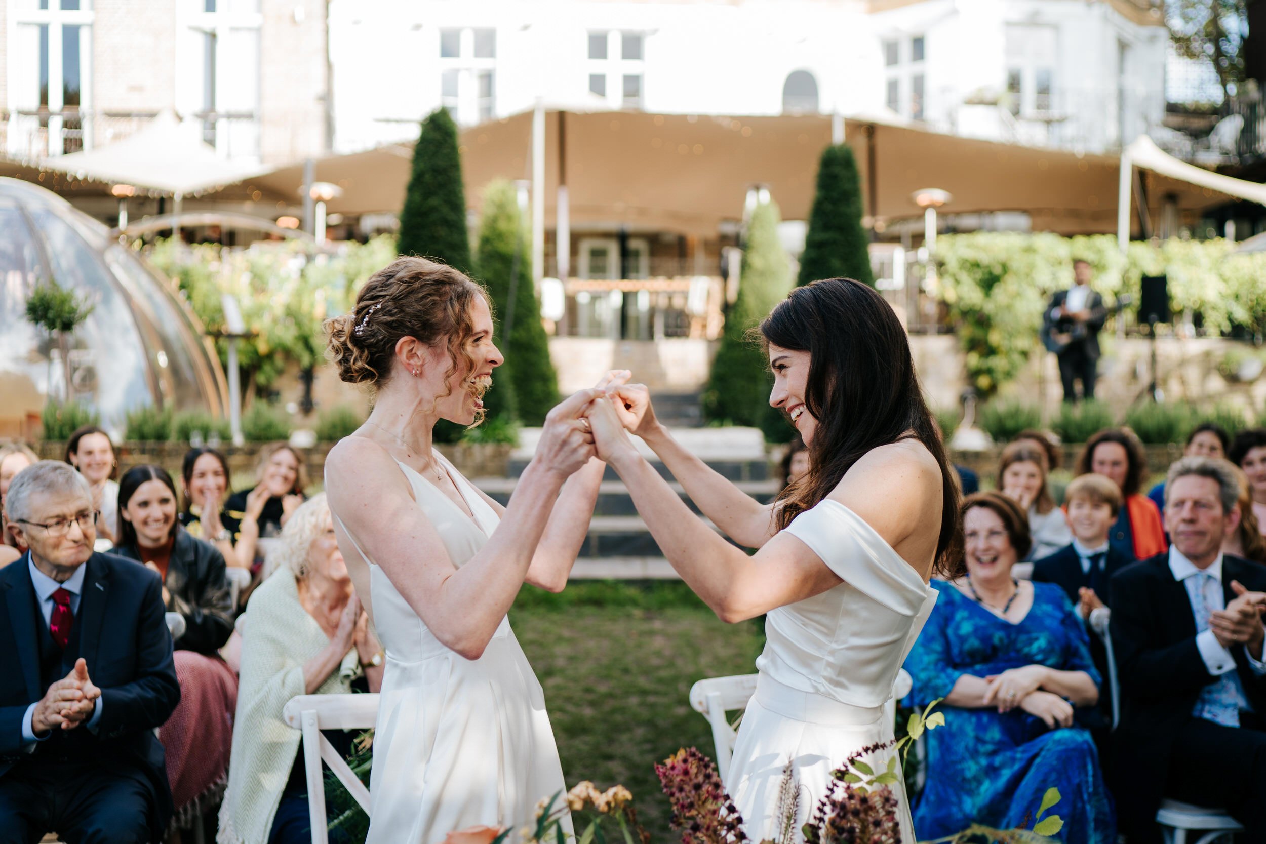 Two brides hold hands and smile at each other at the end of their civil ceremony at Bingham Riverhouse in London